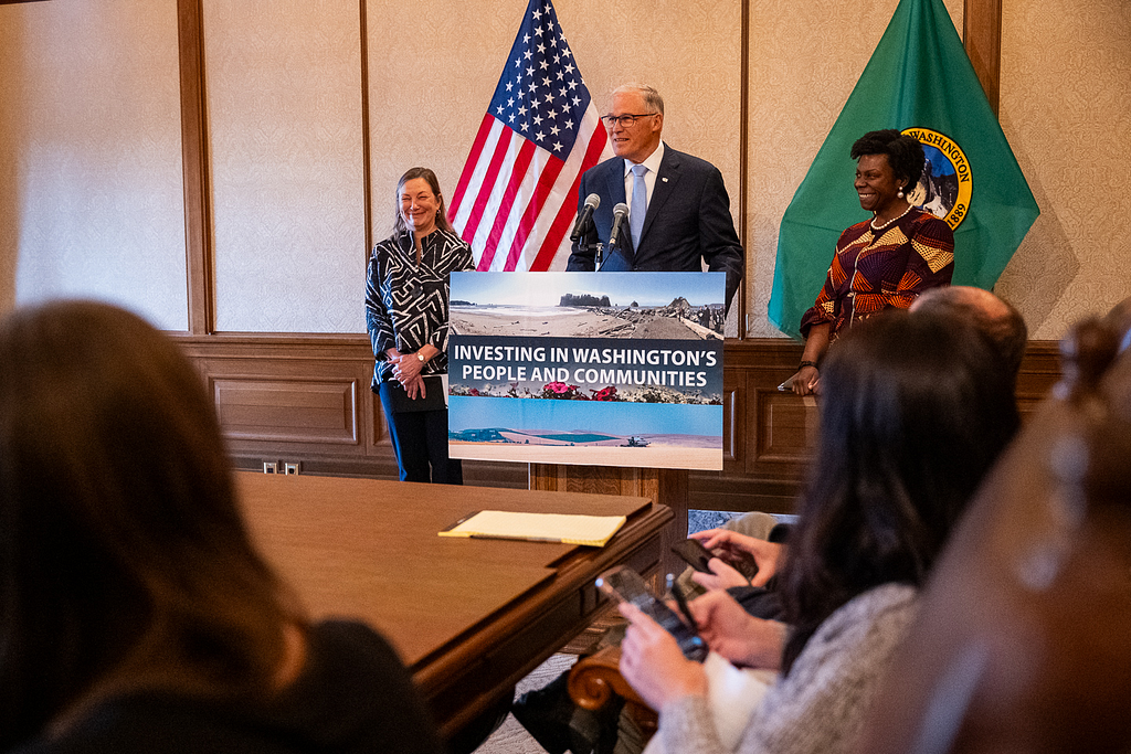 Photo of three people in front of two flags at a podium with a sign: Investing in Washington’s People and Communities