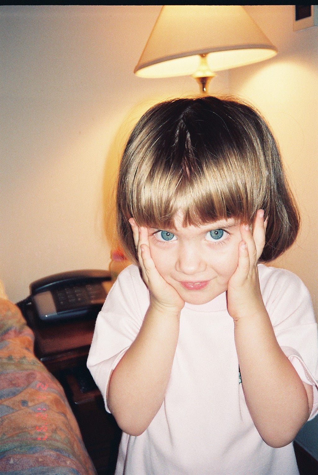 Josefina aged three, touching her face with both her hands. She is wearing a pink top; her hair is short and blonde.