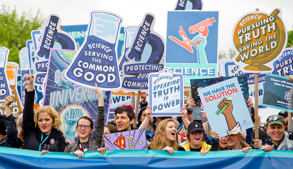 Image of people holding signs saying things like “Science” and “Science Serving the Common Good”