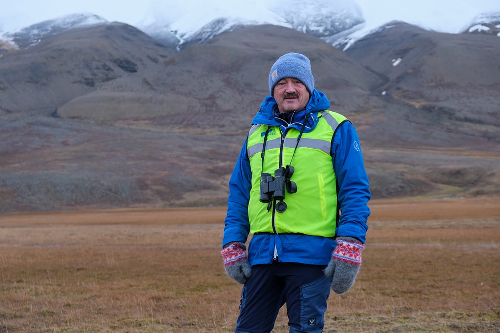 Trond Storm Johansen wearing a bright yellow safety vest over a blue winter jacket, with a pair of binoculars hanging from around his neck. Brown and grey snow-tipped mountains in the backdrop.