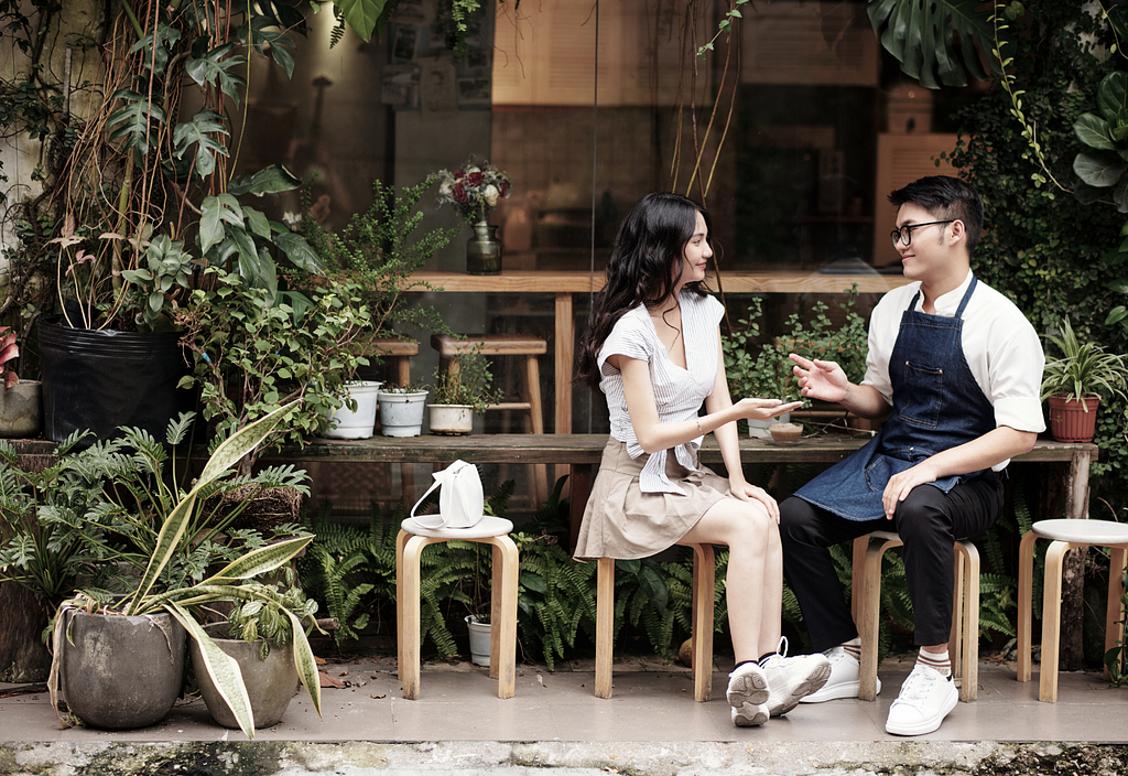 Photo of a young Asian woman chatting with a young Asian man wearing an apron that baristas do, sitting outside a coffee shop and a lot of plans are in front of the shop.