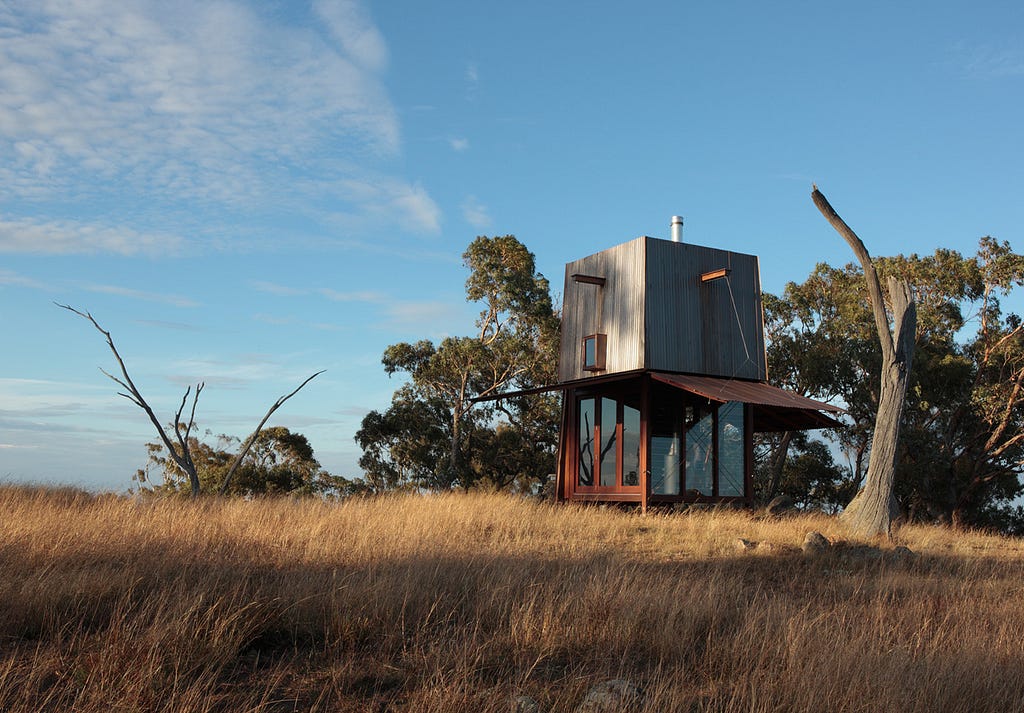 A two storey, tent-like structure sits in a field on the edge of a forest.