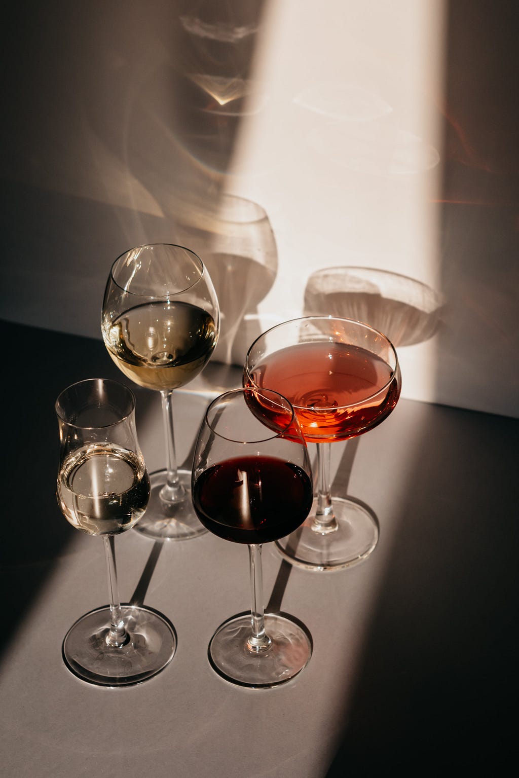 Four clear glass goblets of varying sizes and shapes filled with wine. They are lit by a beam of light as if the light is shining into a dark room from a door being opened.