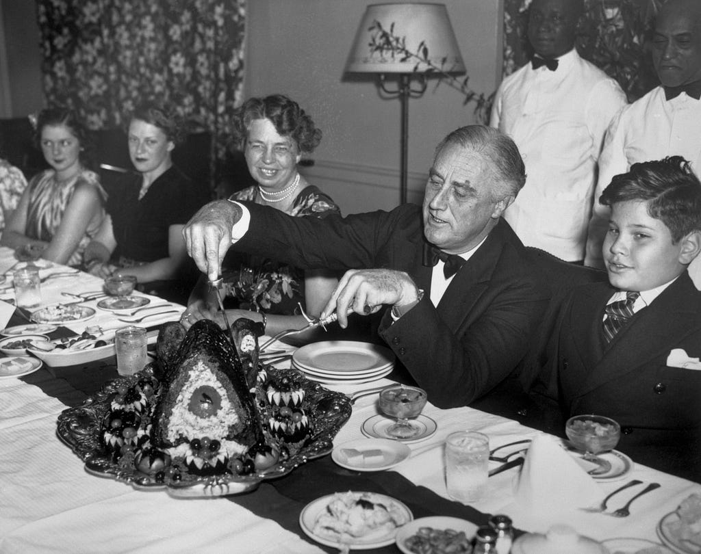 Black-and-white photo of FDR carving a turkey, seated next to Eleanor Roosevelt and some children.