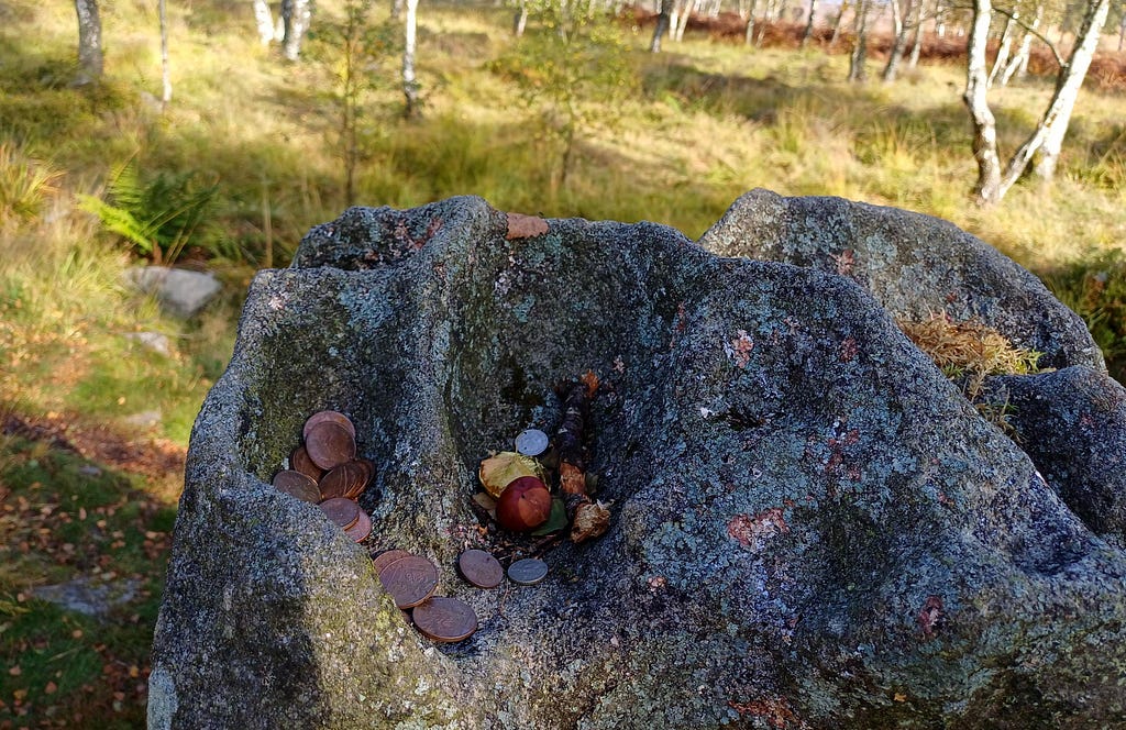 The weather-worn top of a standing stone. The weather has worn holes. Within the holes are copper coins, 2p pieces (UK Stirling currency) left by people passing by.
