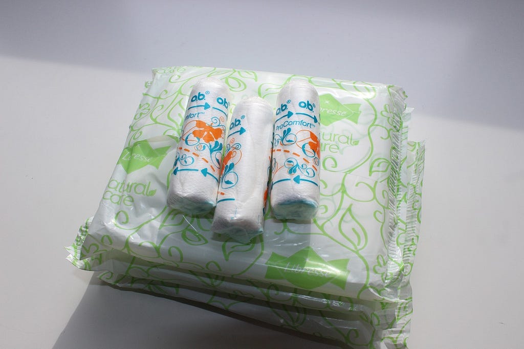 Image shows sanitary pads encased in white plastic with green lines, and on top of that three tampons in their plastic encasings.