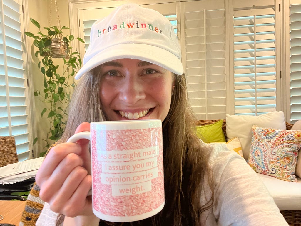 Riesling wearing a white hat with multicolored lettering that says “breadwinner”, and a pink mug that reads “As a straight male, I assure you my opinion carries weight.”