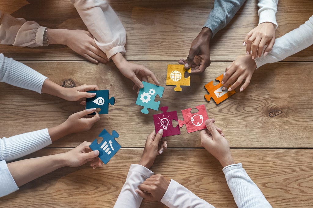 The hands of six people hold differently-coloured jigsaw pieces, representing the building of a complete interdisciplinary picture with different disciplinary elements