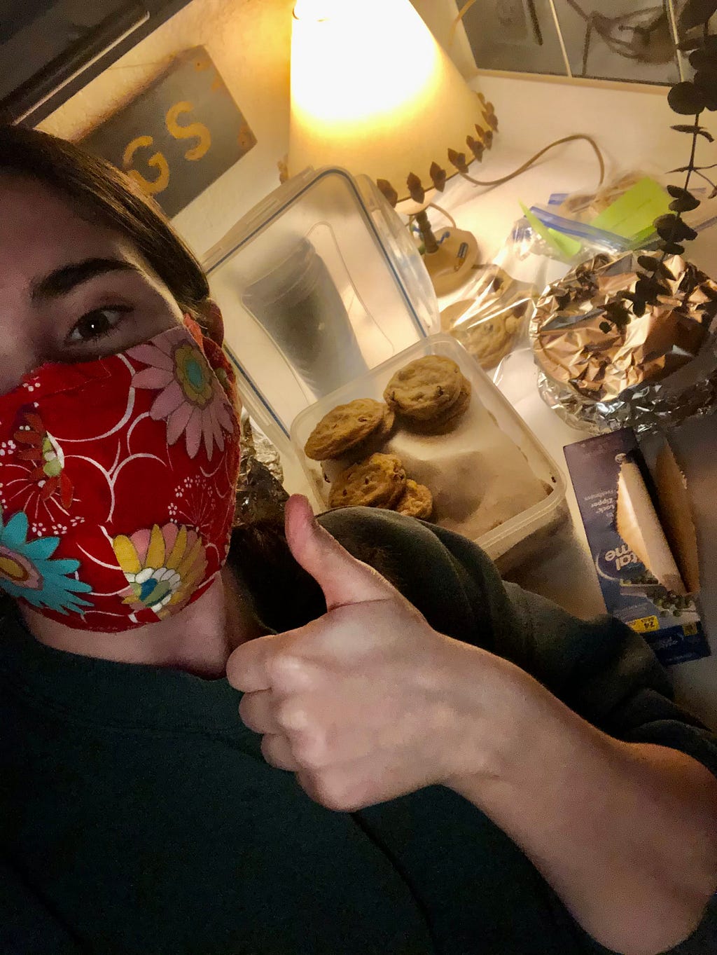 Marney takes a selfie while wearing a red flower print cloth mask. She’s giving a thumbs up to the camera and behind her are baked goods packed into plastic tubs and baggies.