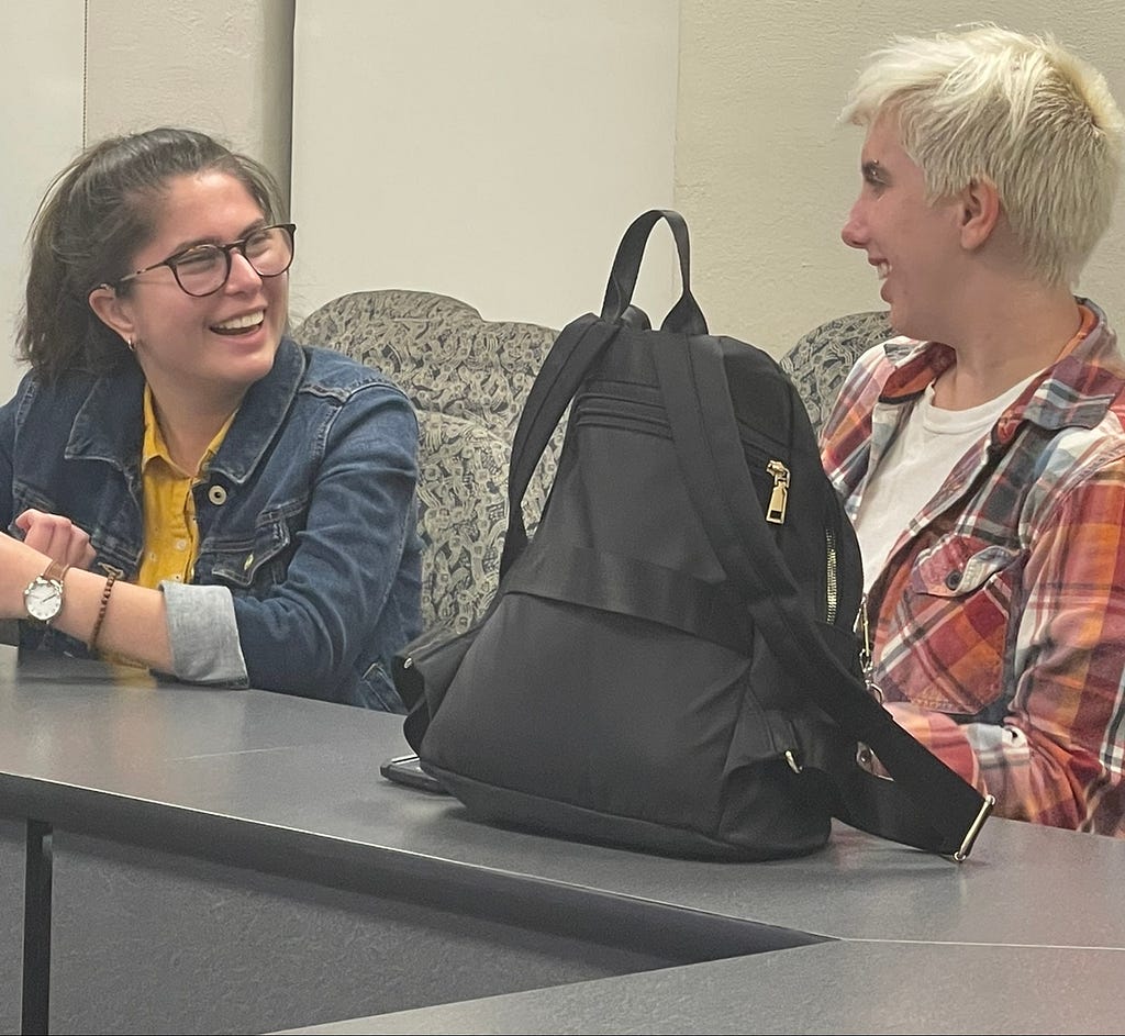 Two students talking to each other in a classroom and one of them is laughing.