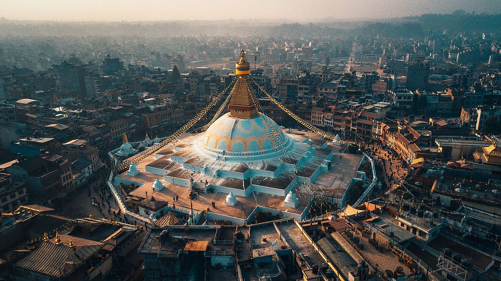 Aerial view of Boudhanath Stupa in Kathmandu, Nepal, during sunset with prayer flags and surrounding buildings.