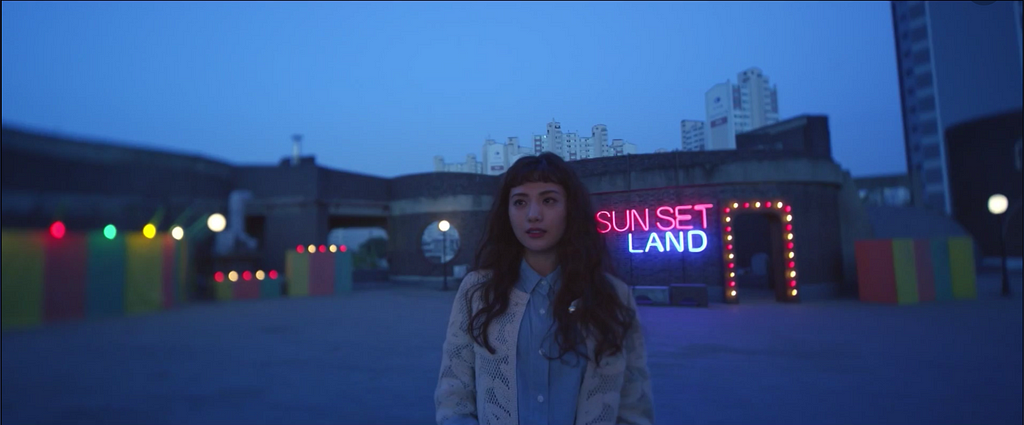 Koo Se-ra stands in front of a neon sign that says Sunset Land.