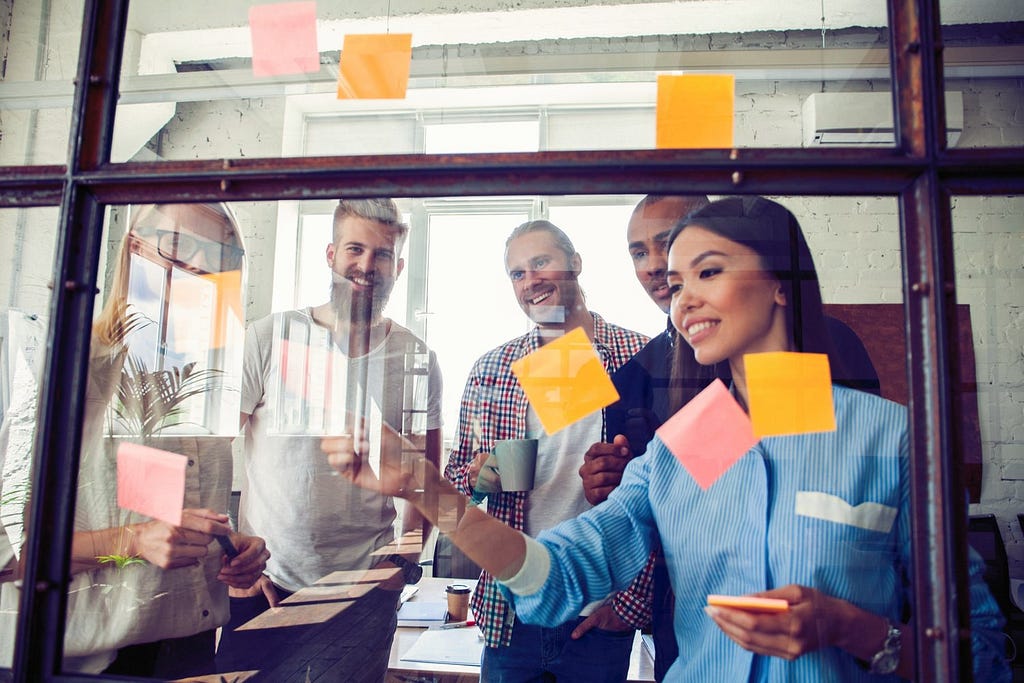 A team of engineers organizes tasks with post-it notes on a window with smiles on their faces now that they have established a quality control system.