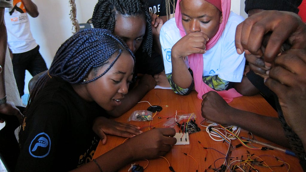 Three African children lean over a table, working on a circuit board, with more people outside the frame.