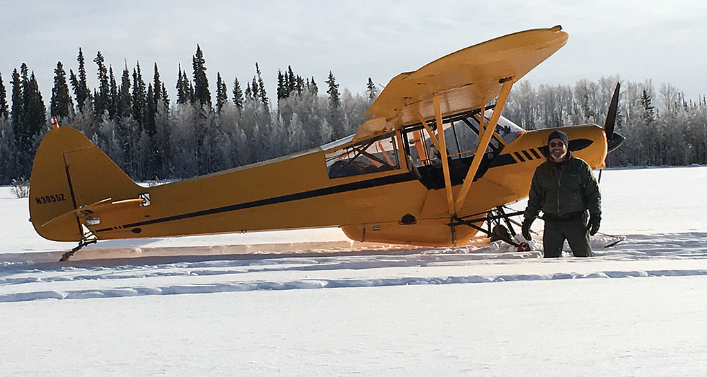 Photo of a yellow airplane on skis in the snow with the pilot.