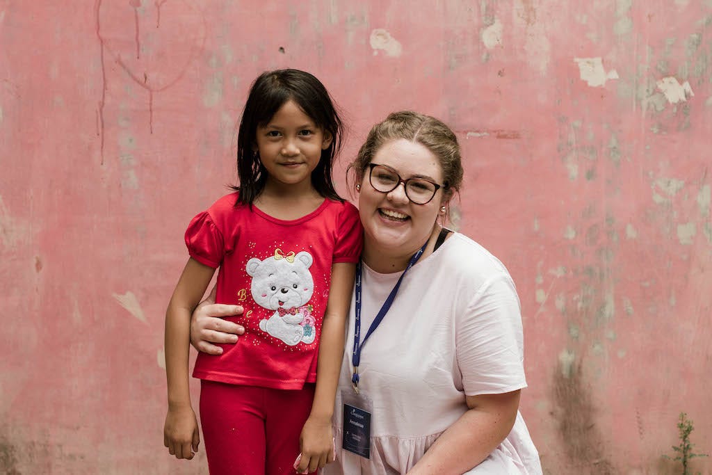 A white sponsor kneels beside an Indonesian girl. Both are smiling and in front of a pink wall.