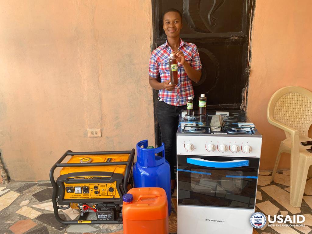 A woman smiles while holding a bottle of castor oil and standing behind new equipment she is using to produce the product.