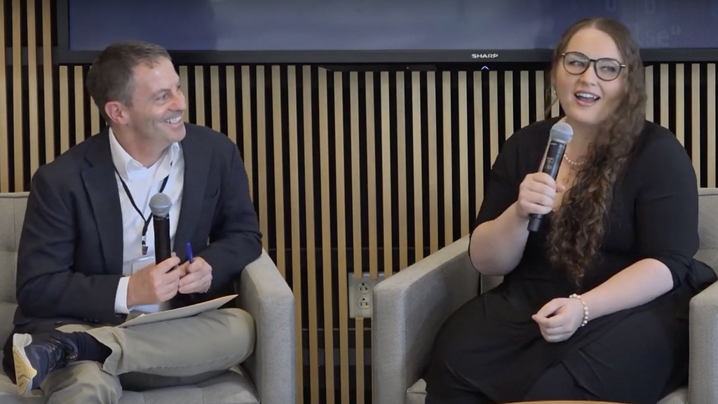 Photo of two adults in business clothes sitting on chairs and speaking into microphones.