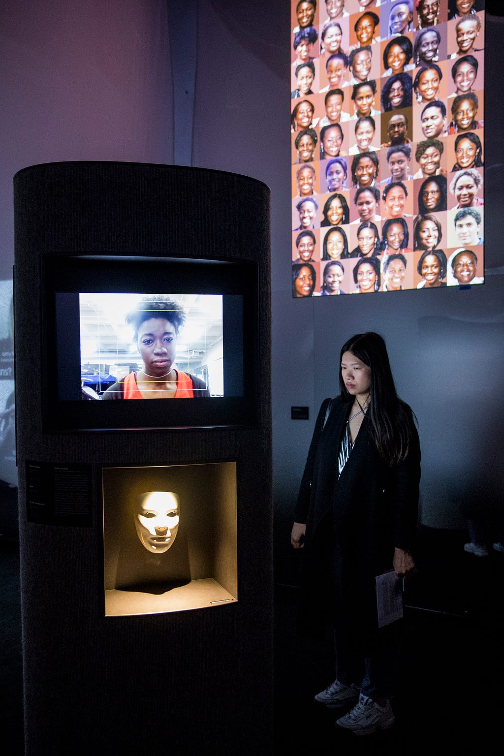 A tall, dark oblong cylindrical plinth containing two niches displaying a video of Joy Buolamwini along with a white mask.