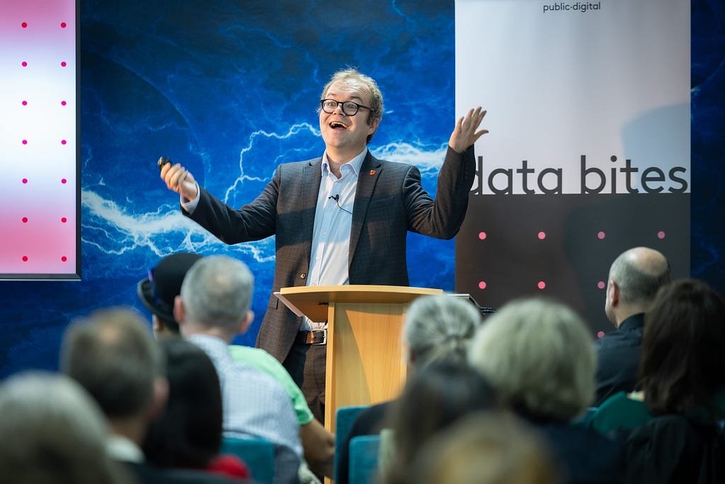 Photo of me, in a suit, stood in front of a lightning background and Data Bites banner, behind a lectern with backs of audience heads visible