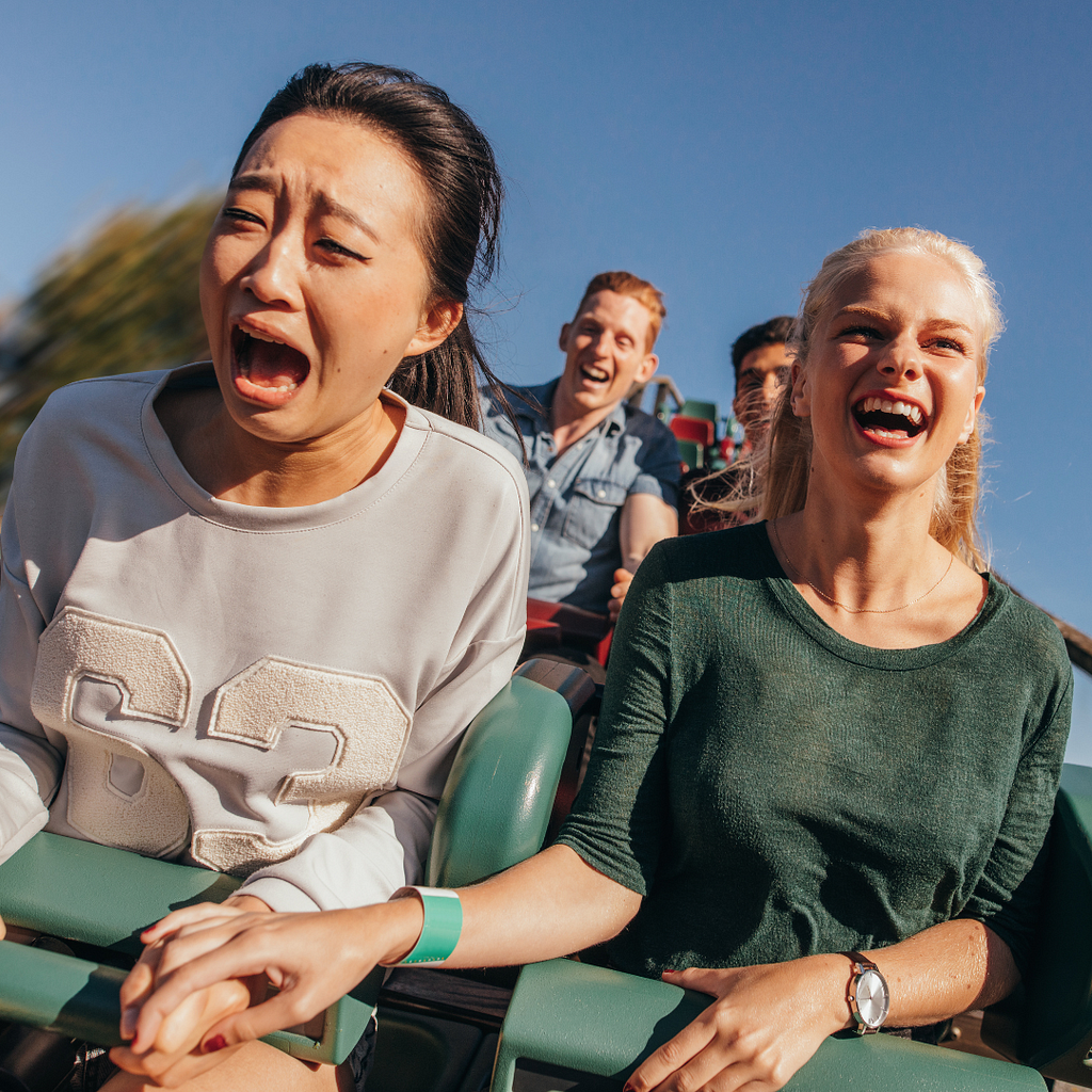 Asian woman and a grey headed white woman hold hands on a rollar coaster and the white woman is laughing and the asian woman is screaming in fear.
