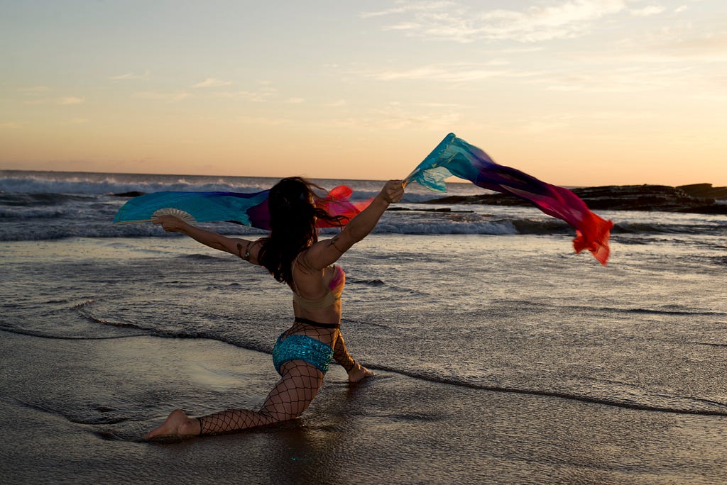 I face away from the camera in a low lunge. In my arms I hold colorful ribbons that dance in the wind. Water pools at my feet and in the distance, waves crash in the sunset.