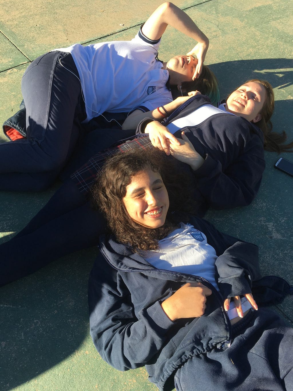 Gianna acostada en el patio de su escuela junto a dos amigues, sonriendo bajo el sol.