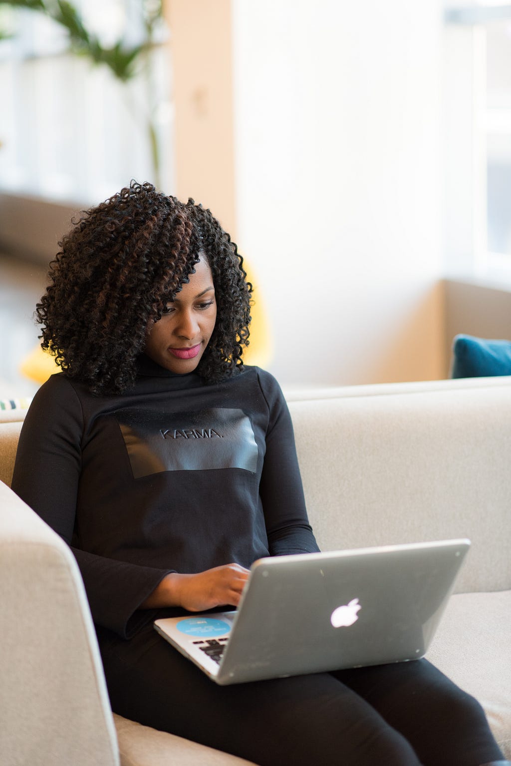 A black woman with curly dark hair wears a long-sleeved black shirt. She is typing on a gray laptop.