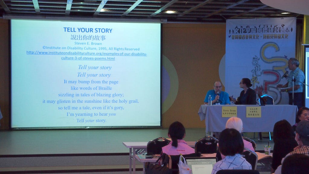 On a stage at a 2016 Conference in Taipei, Taiwan, Steve Brown and Aimée Gramblin sit at a table. To their right is a large screen showing part of the first page of the poem “Tell Your Story.” The backs of several audience members can be seen.