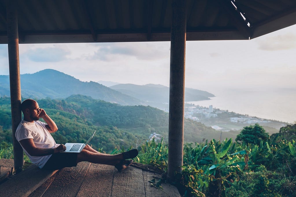 Man sitting with laptop outside, overlooking the sea and mountains