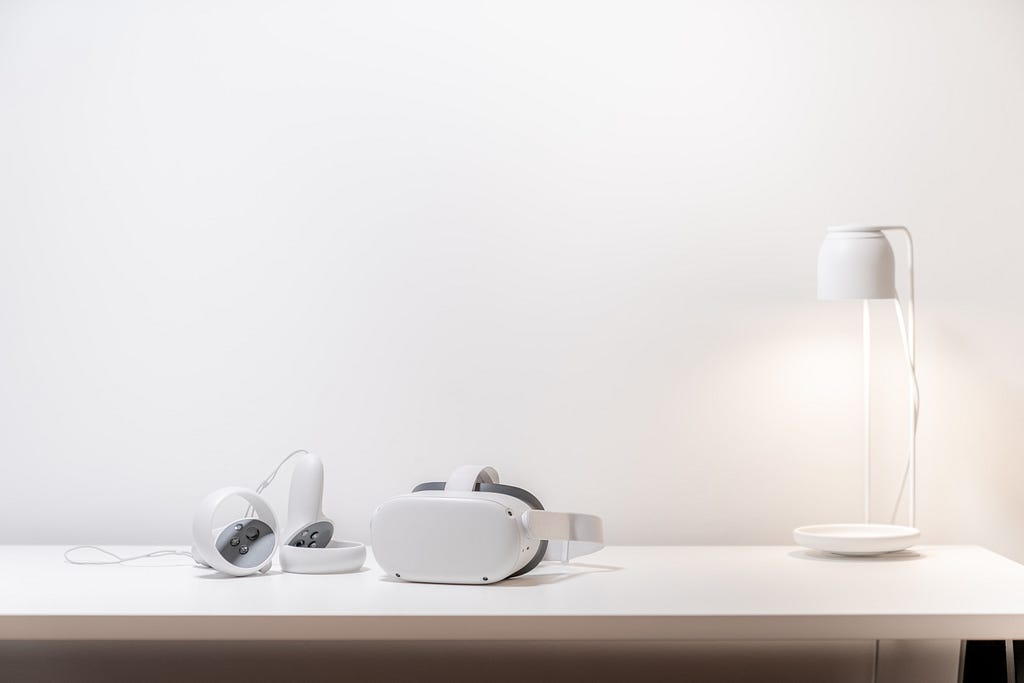 A VR headset and two controllers placed next to a lamp on a desk.