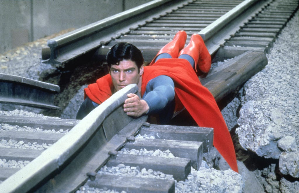 A man dressed in a red cape and boots lying on a broken railway track.