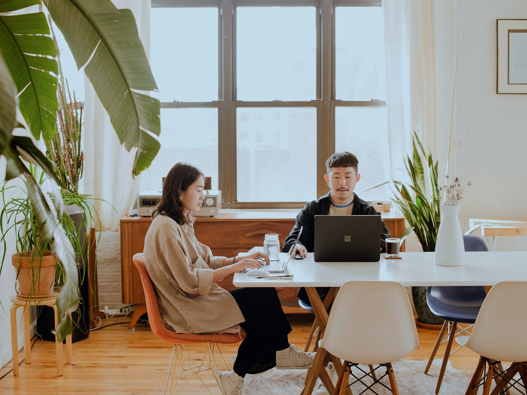 Two humans sitting at a white table with their computers out, surrounded by lush green plants and big windows in a lush office space.