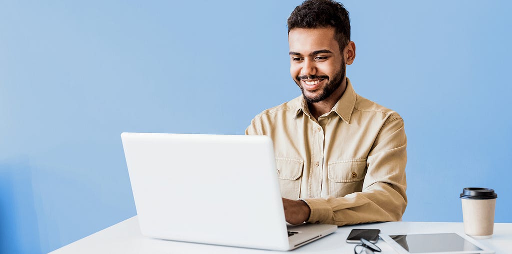 A young man smiling while attending a remote learning experience through a laptop with a blue wall in the background.