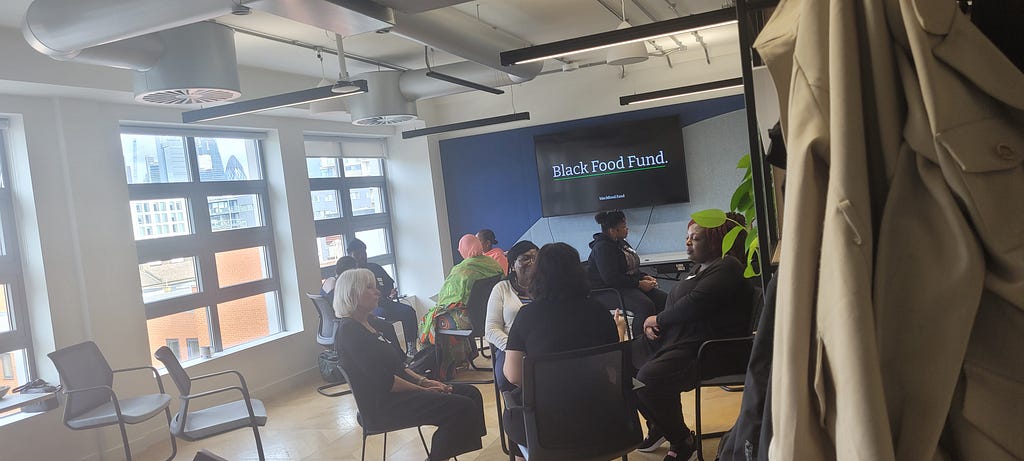 Image of a group of people witting in a room on chairs in a few small cirlces. There is a coat in the foreground and a plant visible. In the background is a screen with the words Black Food Fund. White writing. Black text.