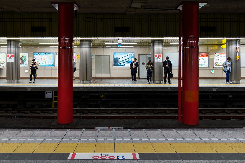 Subway station with red pillars. Men in suits stand looking at their phones waiting for a train to arrive.