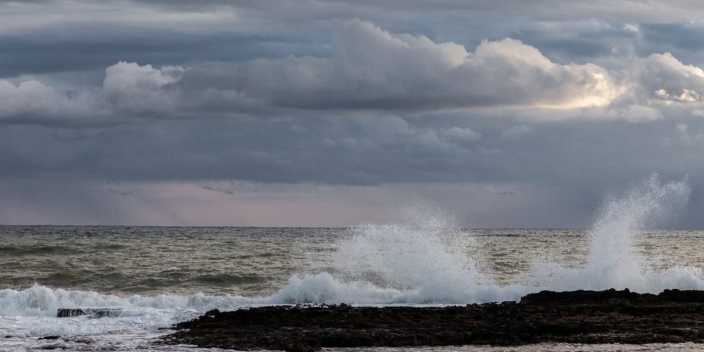 Stormy Mediterranean Sea scene with sea spray hitting against rocks, cloudy sky, silvery gray; Patara, Turkey