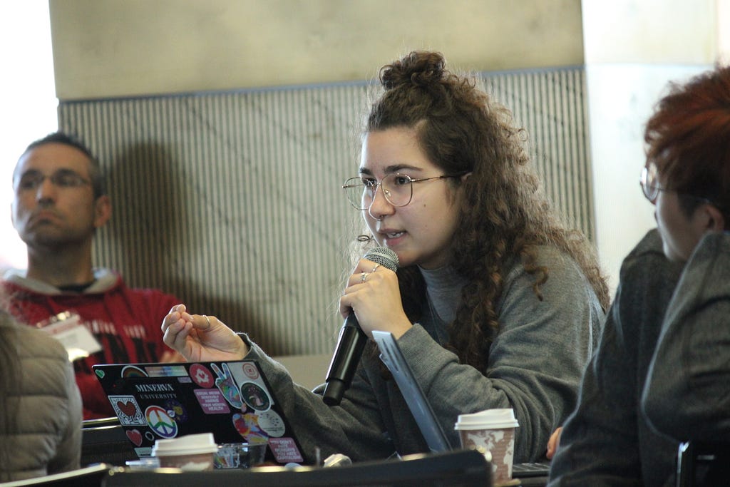 Person sitting in front of a laptop next to other people holding a microphone and asking a question.