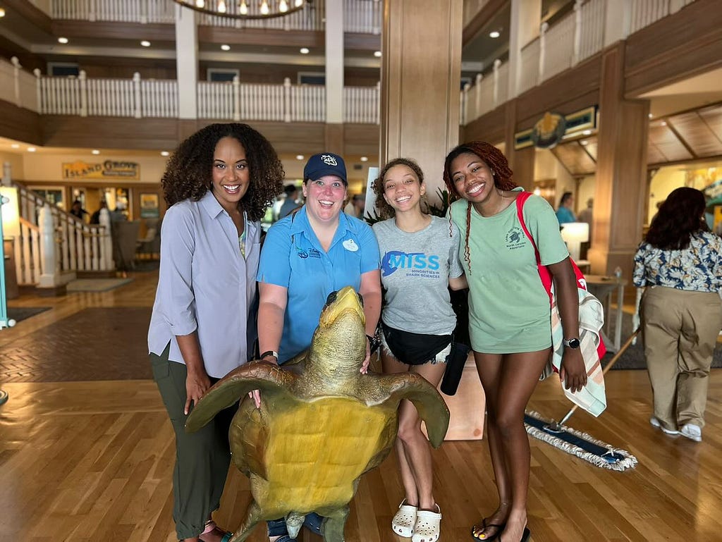 Four people stand indoors around a large sea turtle model. From left to right: one person wears a light-colored button-up shirt with dark pants; the second person wears a short-sleeved blue polo shirt and a baseball cap; the third person wears a gray T‑shirt and casual shorts; and the fourth person wears a light green T‑shirt with a shoulder bag. The space appears to be a public indoor venue with wooden floors, columns, and display signage in the background.