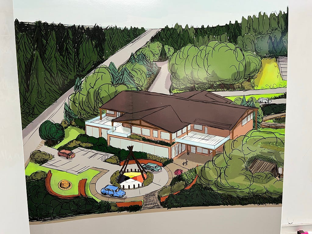 A rendering of the proposed new home of the American Indian Community Center. The brown building is surrounded by green space, parking area and features a colorful storytelling circle in a roundabout.