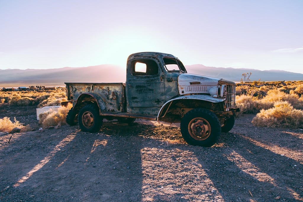 An old style grey single cab truck on scrub land with the low sun behind casting a long shadow.