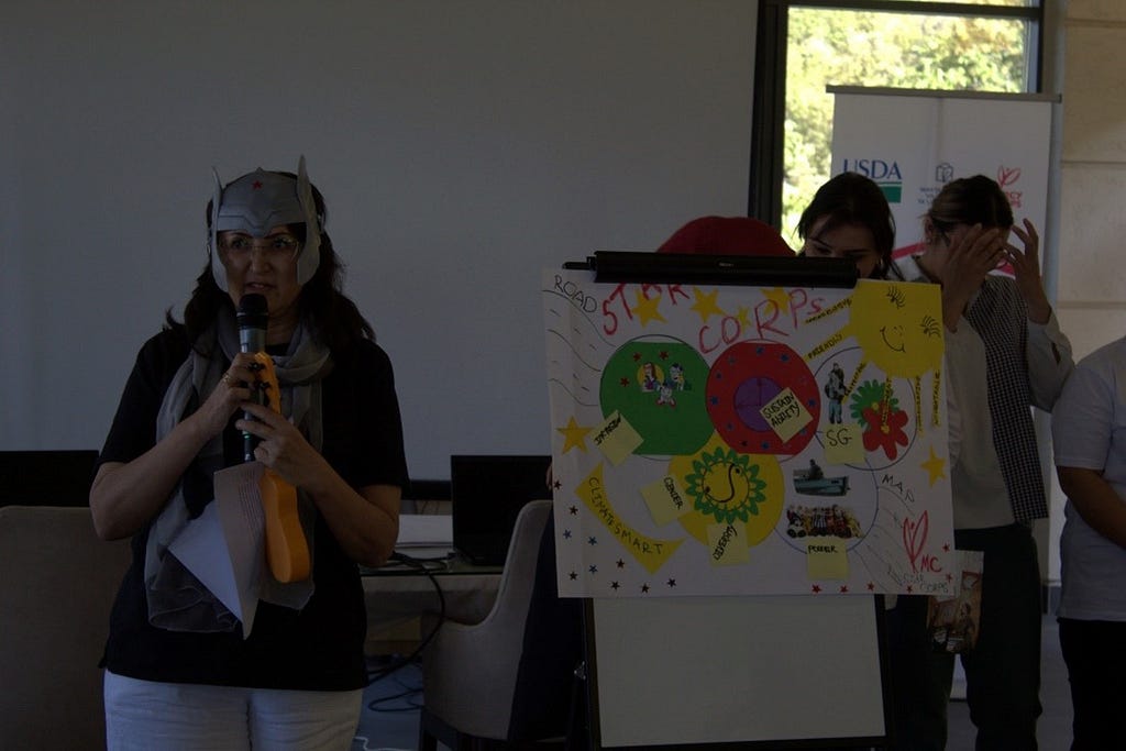A woman wearing a helmet and holding a microphone stands beside an illustration with two people standing behind the paper.