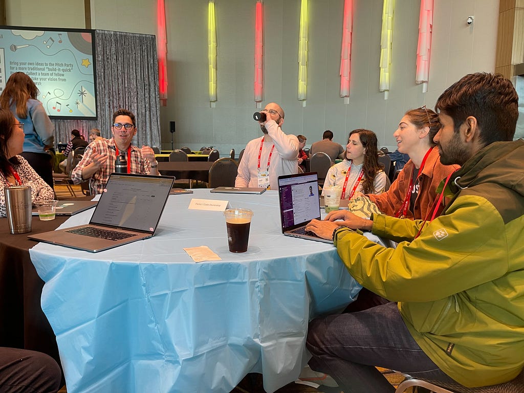 Software developers sitting around a table in a conference hall with laptops open, discussing modularity in large Rails monoliths.