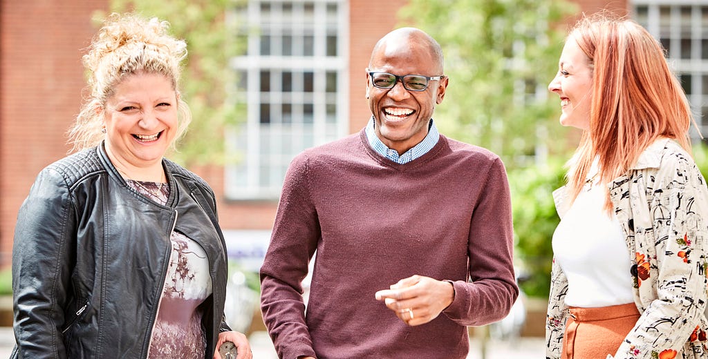 Three mature students — two white women stand either side of a black man who is wearing glasses — laugh together on a university campus.