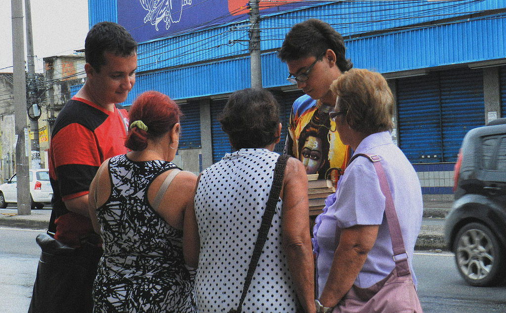 Dois integrantes do coletivo interagem com grupo de três idosas. Avenida e comercial local fechado ao fundo.