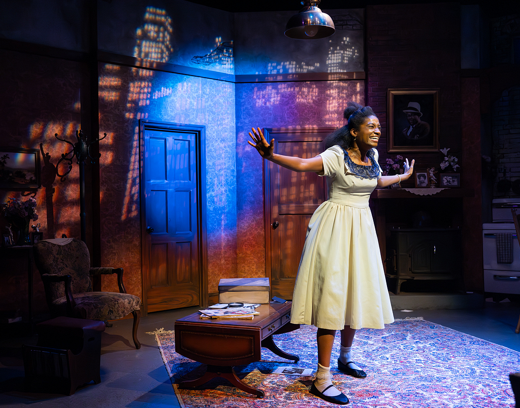 A woman in a white 1950s dress smiles with arms outstretched in front of a cityscape lighting projection in an apartment set onstage.