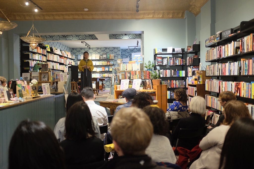 Woman speaking in front of crowd in a cozy bookstore.