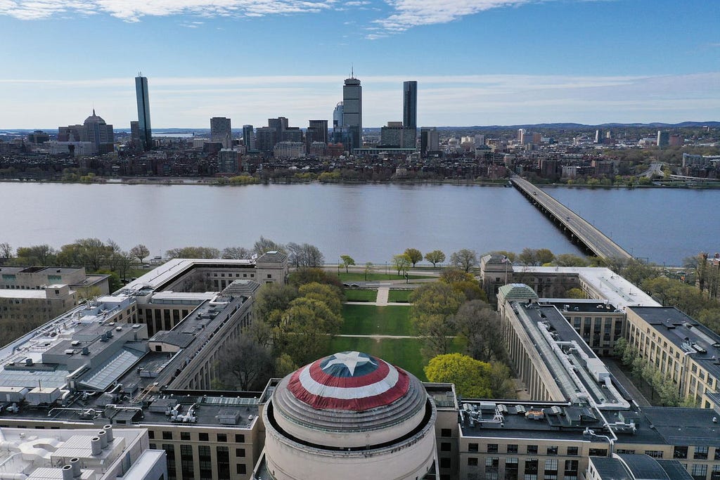 Aerial photo of MIT’s Great Dome with the Captain America shield on it. The view overlooks Killian Court, the Charles River, and the Boston skyline.