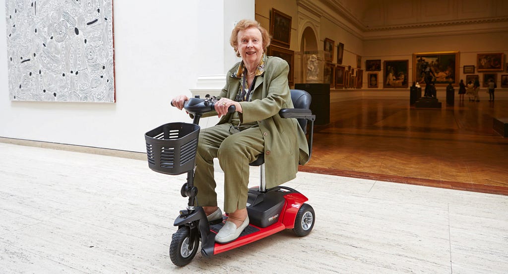 Woman in her 70s is riding a red mobility scooter inside the Art Gallery of New South Wales. She is wearing olive green pants and blazer. She has light skin and red short hair. She is smiling.