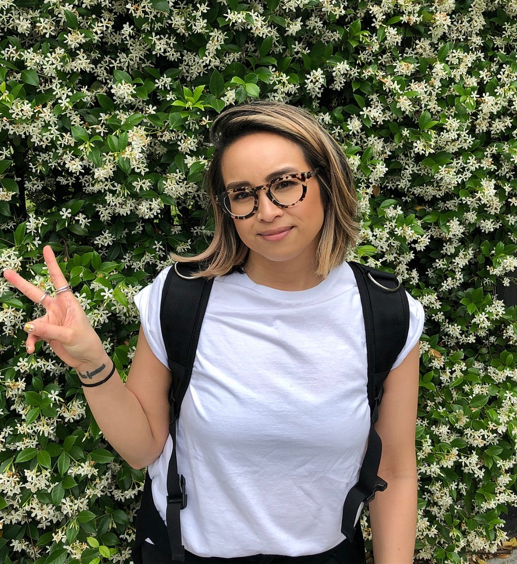 Kat giving the peace sign in front a small, flowering bush.